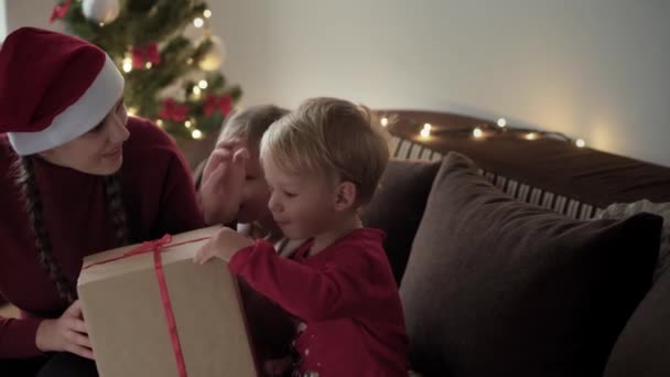 Natal, inverno, ano novo, celebração, conceito de família - Jovem mãe feliz com duas crianças menino e menina vestida com roupas vermelhas caixa de presente aberta sentar perto da árvore de Natal decorada. luz brilhante dele — Vídeo de Stock