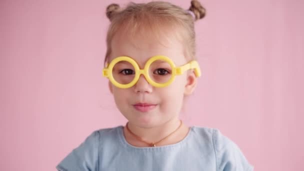 Childhood, celebration, birthday, games concept - close-up little blond girl in looking at camera showing different emotions in yellow toy glasses stretches smile with fingers on pink solid background — Stock Video