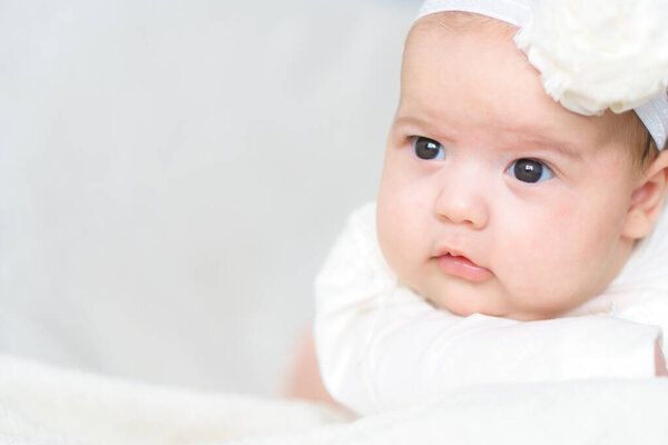 little newborn baby dressed festively in a white dress and a headband lying on his stomach on bed at home. Kind of squinting baby face. Copy space. childhood, development, growth, innocence concepts. High quality photo