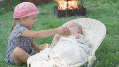 bebeklik, çocukluk, gelişme, yaz tatili konsepti - sallanan sandalyede uzanan ve kız kardeşiyle yeşil çimenlerin piknik arka planında oynayan uyanık bebek portresi. İki çocuk dışarıda.