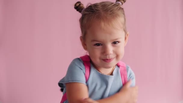 Childhood, school, education, upbringing, science concept - close-up little blonde caucasian slavic girl with backpack flirts closes hands and demonstrates different emotions on solid pink background — Stock Video