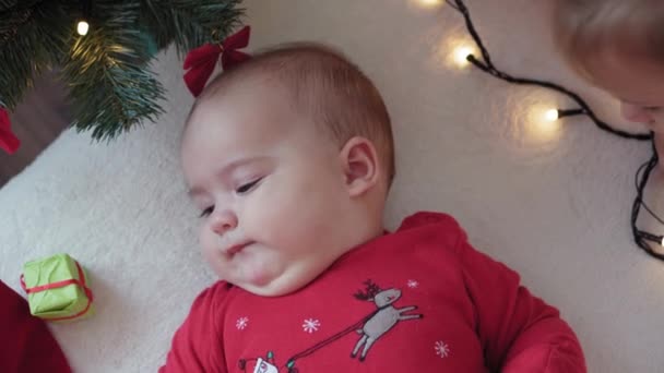 Merry xmas, happy new year, infants, childhood, holidays concept - close-up smiling 6 month old newborn baby in santa claus hat and red bodysuit lay on his back with decorations near christmas tree. — Stock Video