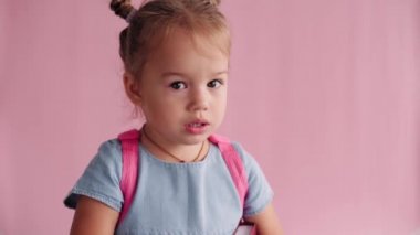 çocukluk, okul, eğitim, yetiştirilme, bilim konsepti - Sırt çantası taşıyan küçük sarışın Slav kız ve katı pembe arka planda hoşnutsuzluk ve anlaşmazlık gösteriyor