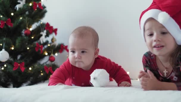 Весёлого Рождества и счастливого Нового года, младенец, детство, праздничная концепция - крупным планом 6-месячный новорожденный младенец в шляпе Санта-Клауса и красном боди-костюме со старшей сестрой ползает с украшениями бал рождественская елка — стоковое видео