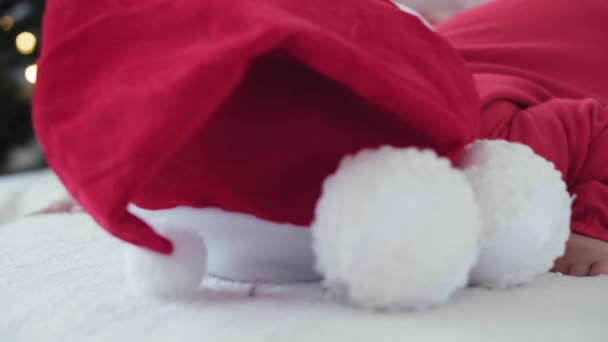 Feliz Navidad y feliz año nuevo, bebés, infancia, concepto de vacaciones - primer plano bebé recién nacido de 6 meses en sombrero de Santa Claus y traje de cuerpo rojo en su barriga se arrastra con bolas de decoración en el árbol de Navidad — Vídeo de stock