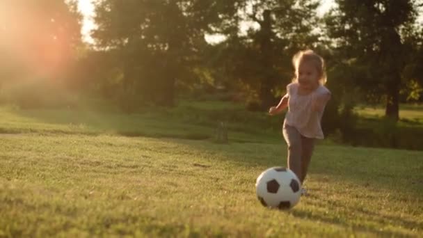 Детство, игры и развлечения, спорт, физкультура, парки и концепция под открытым небом - маленькая блондинка играет в футбол с черно-белым мячом на ярко-зеленой лужайке в летнем парке на закате. — стоковое видео