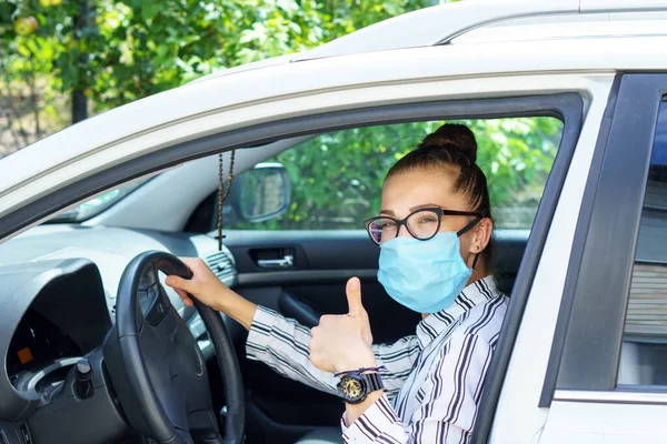 beautiful successful smiling rich business woman in medical protective mask sitting in gray car, wearing glasses showing thumbs up super, prosperous business lady style Lady is driving a car for work.