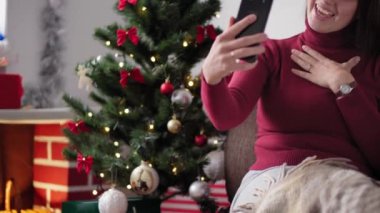 Xmas, kış, yeni yıl, kutlama, aile konsepti - Noel Baba şapkalı mutlu kız, karantinadaki Noel ağacının yanında oturan akıllı telefon dalgalarıyla tatildeki video konferansını uzaktan tebrik ediyor
