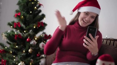 Xmas, kış, yeni yıl, kutlama, aile konsepti - Noel Baba şapkalı mutlu kız, karantinadaki Noel ağacının yanında oturan akıllı telefon dalgalarıyla tatildeki video konferansını uzaktan tebrik ediyor