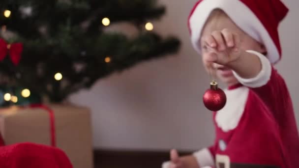 Natal, inverno, ano novo, Celebração, família, Conceito de infância - menino engraçado feliz em chapéu de Santa e roupas de Natal jogar e sorrir com bola vermelha decorar árvore de Natal em férias. — Vídeo de Stock