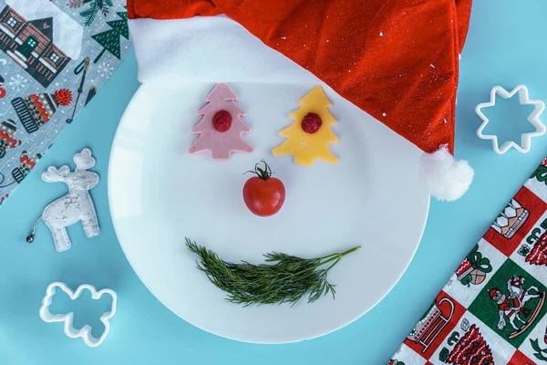 Χριστούγεννα, το χειμώνα, το νέο έτος έννοια - smiley χιονάνθρωπος σε λευκό πιάτο με πιάτα.Μπλε φόντο με Σάντα καπέλο, λουκάνικο τυρί σε σχήμα χριστουγεννιάτικο δέντρο, κόφτες μπισκότων, ελάφι παιχνίδι, κορυφαία θέα, γενικά. Αντιγραφή — Φωτογραφία Αρχείου
