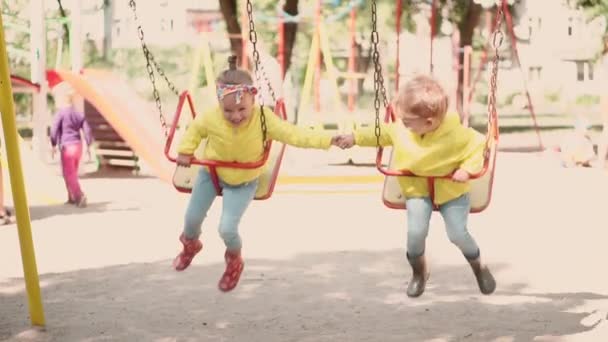 Childhood, game, entertainment, friendship, lifestyle concept - slow motion general plan of two children hold hands in yellow jacket boy and girl 2 and 3 years old joyfully ride on swing on playground — Stock Video