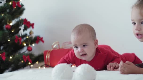Merry xmas and happy new year, infant, childhood, holidays concept - close-up 6 month old newborn baby in santa claus hat and red bodysuit with elder sister crawls with decorations ball christmas tree — Stock Video