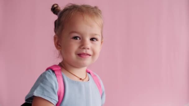 Childhood, school, education, upbringing, science concept - close-up little blonde caucasian slavic girl with backpack demonstrates emotions shows discontent and disagreement on solid pink background — Stock Video