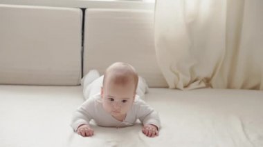 Tıp, sağlık, aile, masumiyet, pediatri, mutluluk, bebek konseptleri - Komik mutlu 7 aylık bebek karnının üstünde emekliyor ve parlak güneş ışığı kreşinde beyaz yumuşak yatakta sallanıyor.