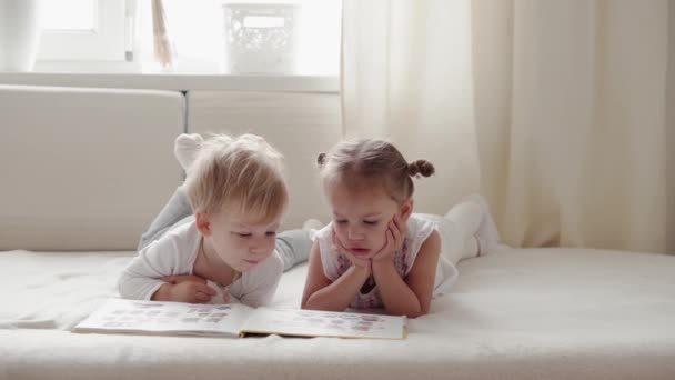 Família, inocência, conceitos infantis - Duas crianças sorridentes ler grande livro interessante de contos de fadas na cama. irmãos menino e menina irmão e irmã se divertir, crianças felizes em quarentena em casa — Vídeo de Stock