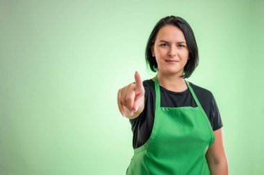 Yeşil önlüklü ve siyah tişörtlü kadın aşçı, yeşil arka planda parmağını göstererek