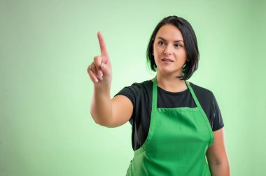 Yeşil önlüklü ve siyah tişörtlü kadın aşçı, yeşil arkaplanda izole edilmiş sanal bir düğmeye basıyor.