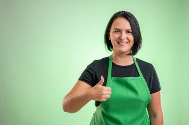 Yeşil önlüklü ve siyah tişörtlü kadın aşçı, yeşil arka planda baş parmaklarını gösteriyor.