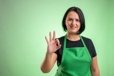 Yeşil önlüklü ve siyah tişörtlü kadın aşçı yeşil arka planda izole edilmiş tamam işareti gösteriyor.