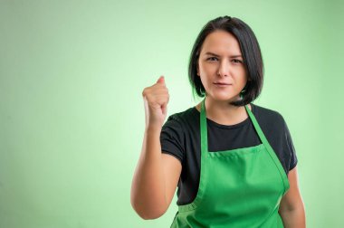 Yeşil önlüklü ve siyah tişörtlü kadın aşçı, yeşil arka planda izole edilmiş yumruğunu gösteriyor.