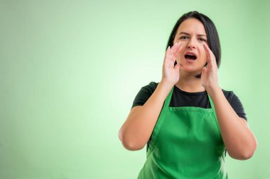 Yeşil önlüklü ve siyah tişörtlü bir kadın aşçı, yeşil arka planda elleri ağzına kadar kapalı bağırıyor.