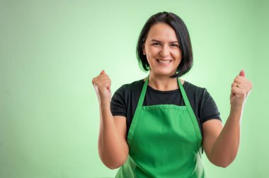 Yeşil önlüklü ve siyah tişörtlü kadın aşçı, yeşil arka planda izole edilmiş neşe ve heyecanla haykırarak yumruklarını kaldırdı.