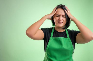 Yeşil önlüklü ve siyah tişörtlü kadın aşçının yeşil arka planda izole edilmiş baş ağrısı var.