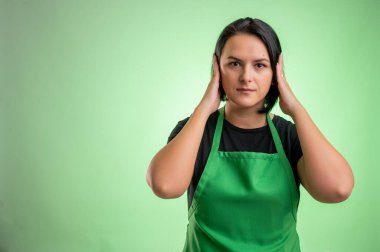 Yeşil önlüklü ve siyah tişörtlü kadın aşçı kulakları sağır kavramı gibi kapatıyor yeşil arka planda izole edilmiş.
