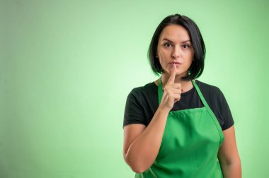 Yeşil önlüklü ve siyah tişörtlü kadın aşçı, yeşil arka planda izole edilmiş sessiz jest gösteriyor.