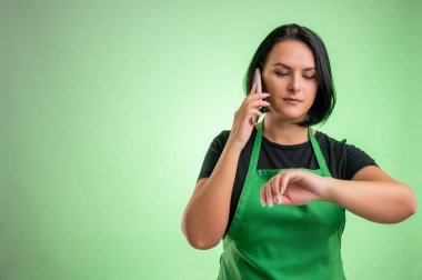 Yeşil önlüklü ve siyah tişörtlü kadın aşçı yeşil arka planda cep telefonuyla konuşuyor.