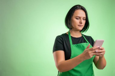 Yeşil önlüklü ve siyah tişörtlü kadın aşçı, elinde akıllı telefon, yeşil arka planda izole edilmiş mesajlar.