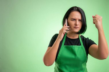 Yeşil önlüklü ve siyah tişörtlü kadın aşçı, hoşnutsuz yüz ve jestler ve yeşil arka planda izole edilmiş cep telefonuyla konuşma.