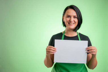 Yeşil önlüklü ve siyah tişörtlü kadın aşçı, yeşil arka planda izole edilmiş beyaz bir kağıt tutuyor.