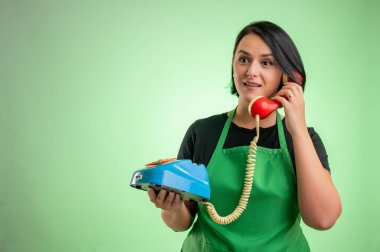 Yeşil önlüklü ve siyah tişörtlü kadın aşçı yeşil arka planda izole edilmiş eski bir telefonla konuşuyor.