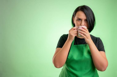 Yeşil önlüklü ve siyah tişörtlü bir kadın aşçı yeşil arka planda izole edilmiş bir kafe içiyor.