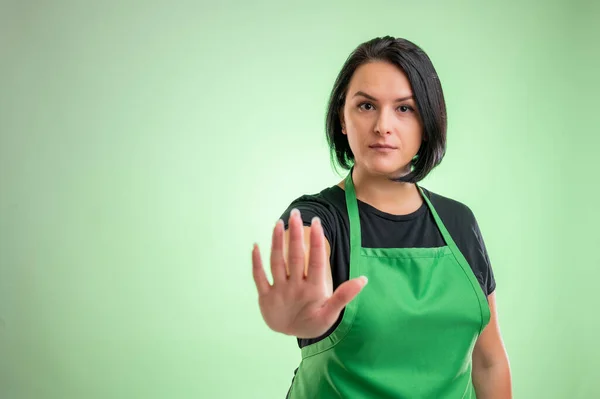 Female cook with green apron and black t-shirt, showing stop sign isolated on green background