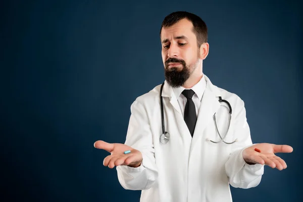 手の上に赤と青の丸薬を示す医学の制服で聴診器を持つ男性医師の肖像 疑いの青い隔離された背景にポーズ — ストック写真