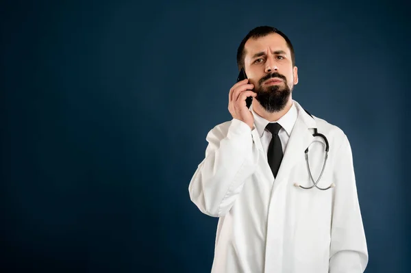 Portrait of male doctor with stethoscope in medical uniform talking on cellphone, smartphone posing on a blue isolated background.