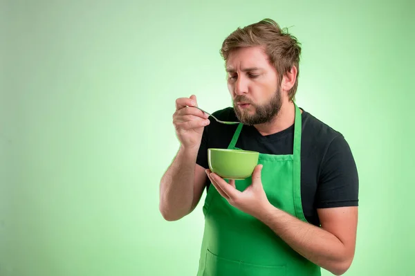 Supermarket employee with green apron and black t-shirt, cool the soup, hold the bowl in his hand  isolated on green background