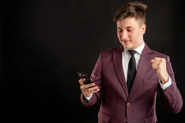 Law student with blond hair dressed in burgundy jacket, white shirt and black tie reading and sending text messages on cellphones happy face posing on isolated black background with copy space advertising area