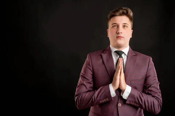 Law student with blond hair dressed in burgundy jacket, white shirt and black tie praying posing on isolated black background with copy space advertising area
