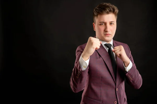 Law student with blond hair dressed in burgundy jacket, white shirt and black tie showing fists, boxing posing on isolated black background with copy space advertising area