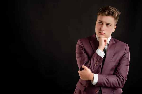 Law student with blond hair dressed in burgundy jacket, white shirt and black tie holding hand under his chin, having doubtful posing on isolated black background with copy space advertising area