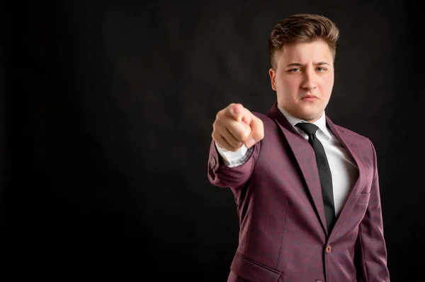 Law student with blond hair dressed in burgundy jacket, white shirt and black tie with angry face pointing his finger posing on isolated black background with copy space advertising area
