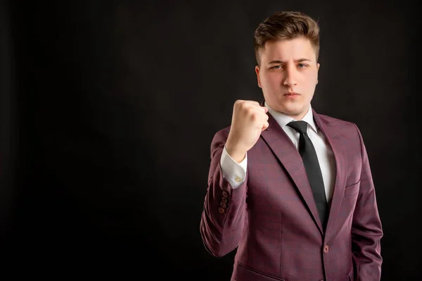 Law student with blond hair dressed in burgundy jacket, white shirt and black tie showing fist posing on isolated black background with copy space advertising area