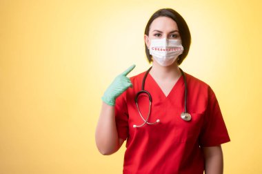 Steteskoplu güzel bir kadın doktor portresi kırmızı önlük giyiyor, koruyucu bir maske takıyor, parmakları sarı izole bir arka planda poz veriyor..