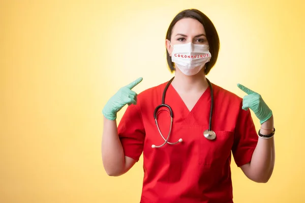 Portrait of beautiful woman doctor with stethoscope wearing red scrubs, wears a protective mask, with her fingers pointed posing on a yellow isolated background.