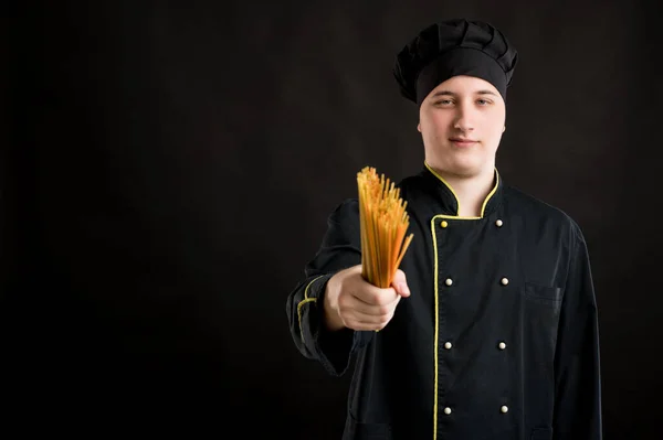 Young male dressed in a black chef suit shows spaghetti posing on a black isolated background with copy space advertising area