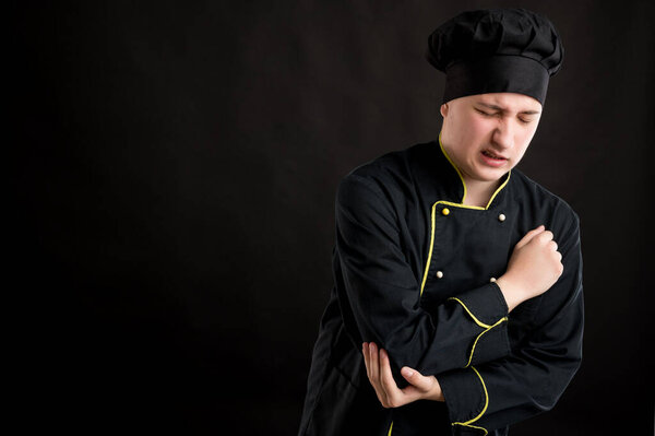 Portrait of young male dressed in a black chef suit elbow pain posing on a black isolated background with copy space advertising area